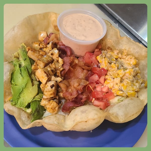 A taco salad in a crispy tortilla bowl with chicken, bacon, avocado, corn, tomatoes, and dressing.